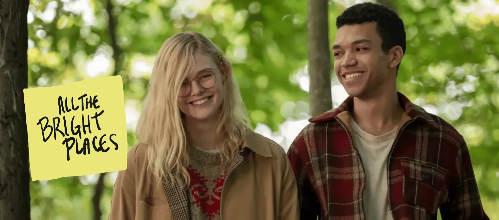 A medium shot set outdoors against a lush, out-of-focus backdrop of green trees, featuring lead actors Elle Fanning and Justice Smith from the 2020 film All the Bright Places walking side by side and smiling broadly. On the left, Elle Fanning, playing Violet Markey, has long blonde hair parted on the side, round glasses, and wears a tan coat over a cozy patterned sweater with red details. To her right, Justice Smith, playing Theodore Finch, looks toward her with a warm expression, wearing a red-and-black plaid flannel jacket over a neutral t-shirt. On the left foreground, attached to a tree trunk, is a bright yellow sticky note graphic featuring handwritten black text that reads "ALL THE BRIGHT PLACES."