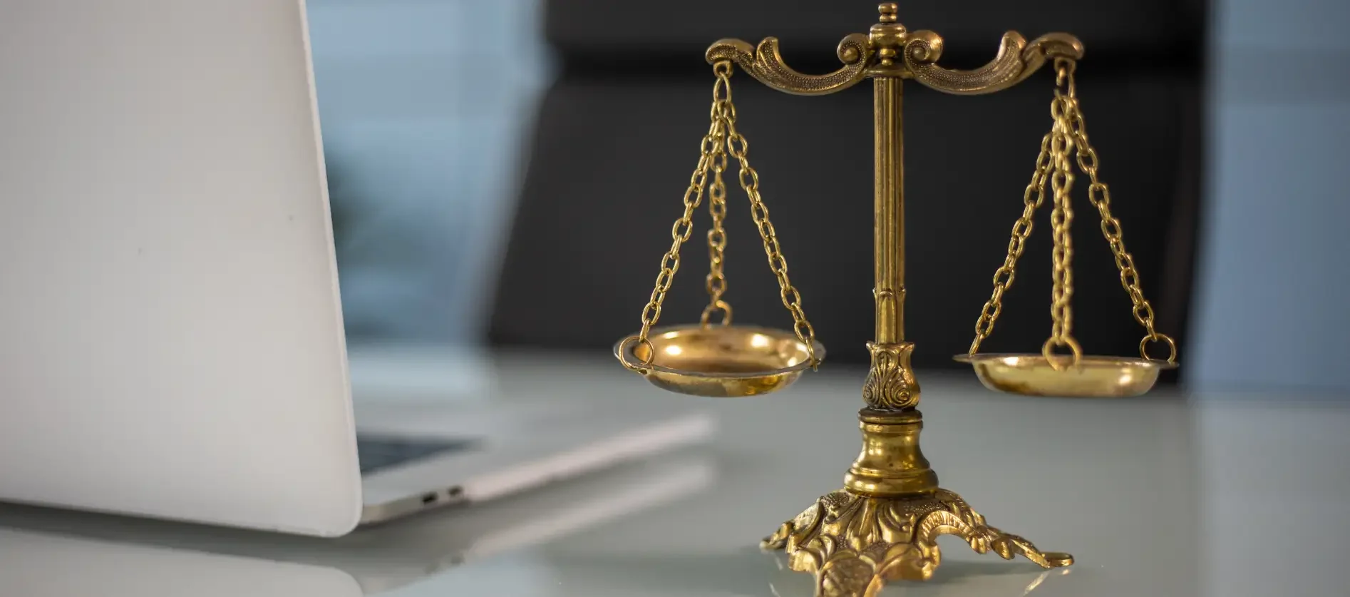 An ornate, vintage brass scale of justice sits on a clear glass tabletop inside a bright modern office. The scale is intricately detailed with a decorative base, a center post, and two balance pans suspended by chains. To the left, a sleek white laptop rests open, partially out of frame, juxtaposing classical legal symbolism with modern technology against a soft-focus background.