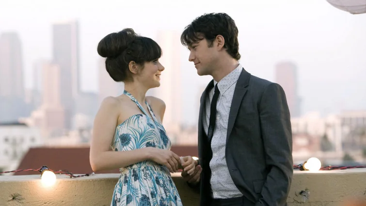A medium shot captures actors Zooey Deschanel and Joseph Gordon-Levitt standing close together during an outdoor rooftop gathering at dusk, set against a hazy, soft-focus urban skyline. Zooey Deschanel stands on the left, looking up smilingly into his eyes with her hair styled in an elegant, voluminous high bun with thick bangs. She is wearing a sleeveless halter dress featuring a white background adorned with a blue botanical and floral pattern. Joseph Gordon-Levitt stands on the right, facing her with a gentle, reciprocating smile. He is dressed in a dark gray tailored suit jacket over a light gray, subtly patterned button-up shirt and a slim black tie. In the foreground, the edge of a concrete rooftop ledge is visible, decorated with a string of glowing, warm-toned bulb lights. The background features the blurred silhouettes of city skyscrapers under a pale, overcast sky.