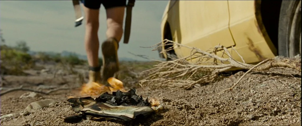ground-level cinematic frame, the lower half of a traveler, Christopher McCandless, is seen from behind as they walk away down a dirt path. The individual is wearing dark shorts and sturdy hiking boots, with heavy straps from a backpack hanging down beside their legs. To the right, the front side panel, tire, and wheel well of a dusty, faded yellow sedan are parked askew on the rocky ground. In the immediate foreground, a small stack of paper or paper money burns on the dirt, flames licking at the charred edges. The environment is harsh and arid, filled with loose gravel, dry soil, and a brittle, dead branch resting near the car's tire. In the background, the horizon stretches out under a hazy, pale sky with faint, distant mountains fading into the heat.