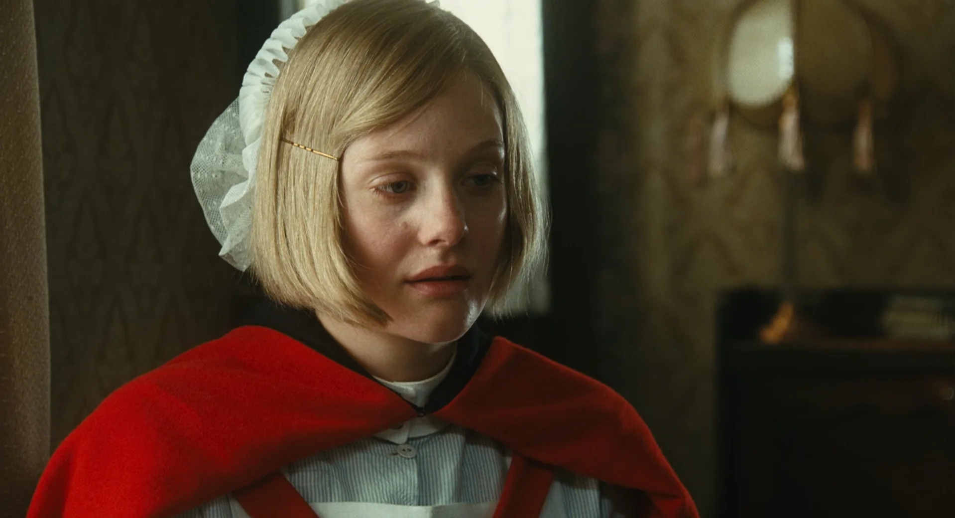 An emotional close-up captures a young blonde woman from the chest up, her tear-filled, glassy eyes gazing down in deep sorrow. Her short bob is styled with a side-swept fringe held by a simple gold hairpin, and a delicate white lace bonnet rests at the back of her head. She wears a crisp, light blue collared blouse underneath a bright red cape draped over her shoulders. The muted lighting casts a somber mood, illuminating her pale complexion and flushing her cheeks and nose. In the softly blurred background, a patterned wallpapered room features a vintage lamp with hanging tassels, accentuating the historical or period setting.