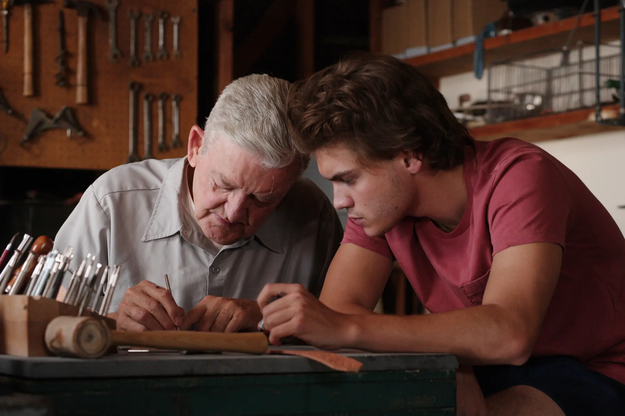 Hal Holbrook and Emile Hirsch as Ron Franz and Christopher McCandless with gray hair and a younger man with brown hair lean closely over a green wooden workbench in a well-lit workshop. The older man, wearing a light gray short-sleeved button-down shirt, carefully uses a tool on a piece of leather while the younger man, in a reddish-pink t-shirt, watches intently. In the foreground, a wooden block holds a collection of carving and stamping tools alongside a wooden mallet. The background features a tool organization board mounted on the wall, filled with wrenches and hammers, as well as shelves stocked with storage containers.