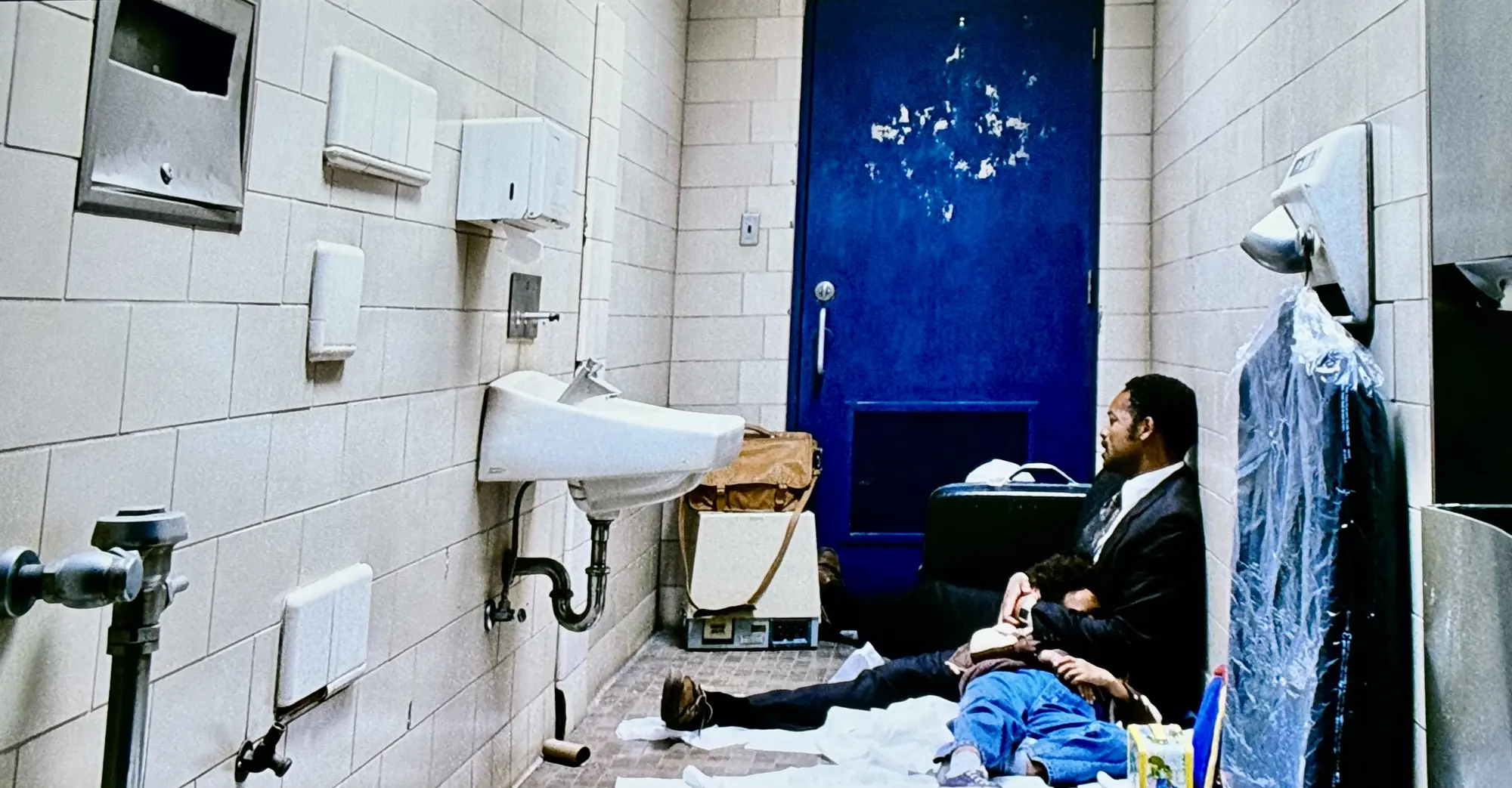 A wide shot depicts a scene from a film set inside a narrow, stark public restroom with light beige tiled walls. A dark blue metal door with peeling paint stands at the far end of the room. On the left wall, there is a white porcelain sink with exposed plumbing, alongside various fixtures like hand dryers and soap dispensers. On the right, a man in a black suit jacket and dark trousers sits on the floor with his back against the wall, holding a young child wearing blue pants who is sleeping in his lap. A large leather duffel bag rests on a machine next to the sink, and a black suitcase sits behind the man. To the right of the man, a suit jacket wrapped in a clear plastic garment bag hangs from a wall fixture. The floor around them is scattered with paper towels and personal items.