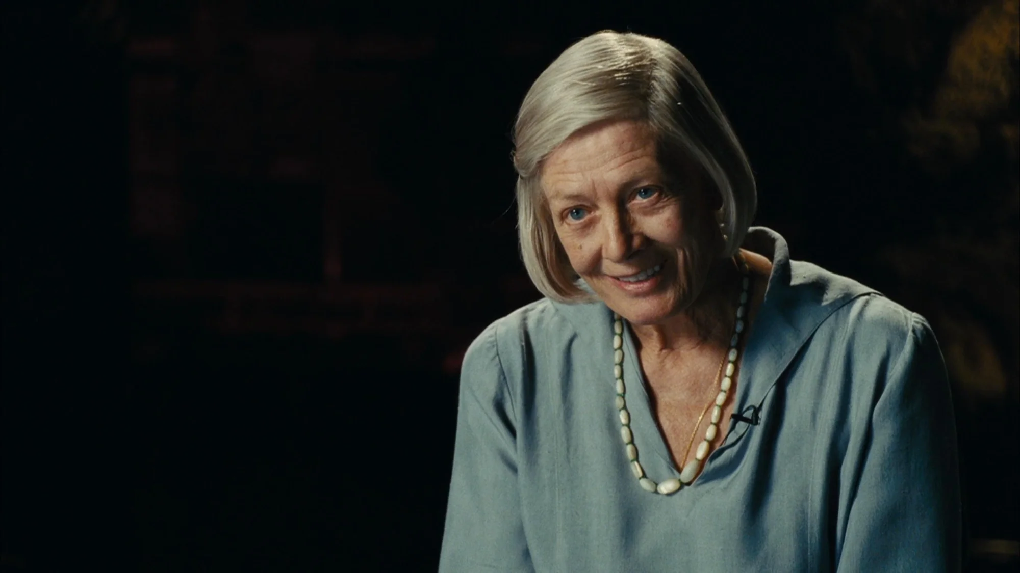 An older woman with short, chin-length grey hair, parted on the side, looks toward the camera with a gentle, warm smile. She is wearing a light blue, collared blouse with an attached lavalier microphone clipped to the left side of the collar. Around her neck is a long, prominent necklace made of light-colored, elongated oval beads, layered over a thin gold chain. The lighting highlights her face, silver hair, and the texture of her skin, while the background remains completely dark and out of focus.