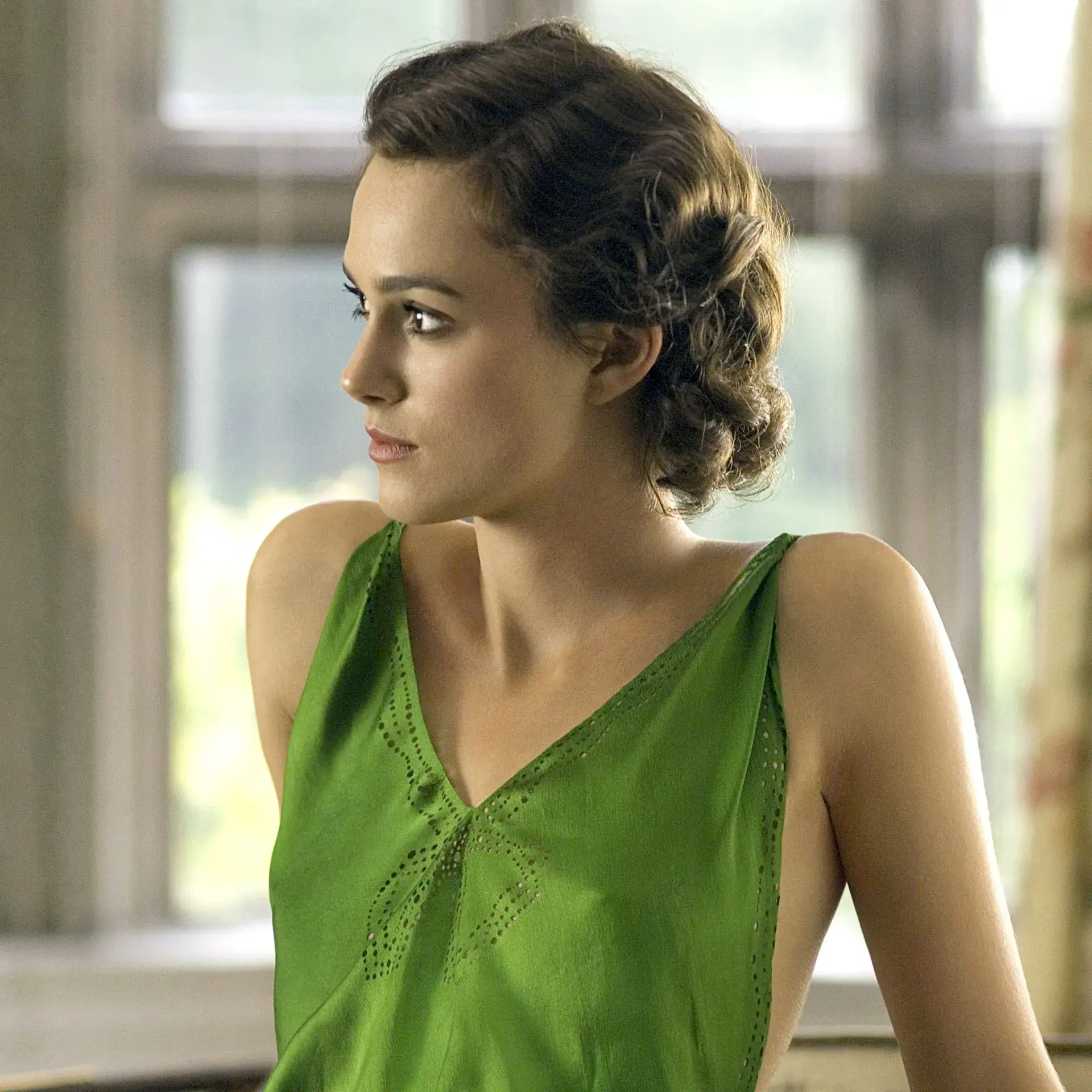 A medium profile shot of a woman with light brown skin, dark brown hair, and sharp facial features, looking slightly to the left. Her hair is styled in structured 1930s-style waves and gathered at the back. She wears a vibrant, bright green silk or satin sleeveless dress featuring a deep V-neckline adorned with delicate, patterned eyelet cutouts or embroidery across the bodice. The background shows a softly blurred, brightly lit room with large grid-patterned windows and a hint of a floral curtain on the right side.
