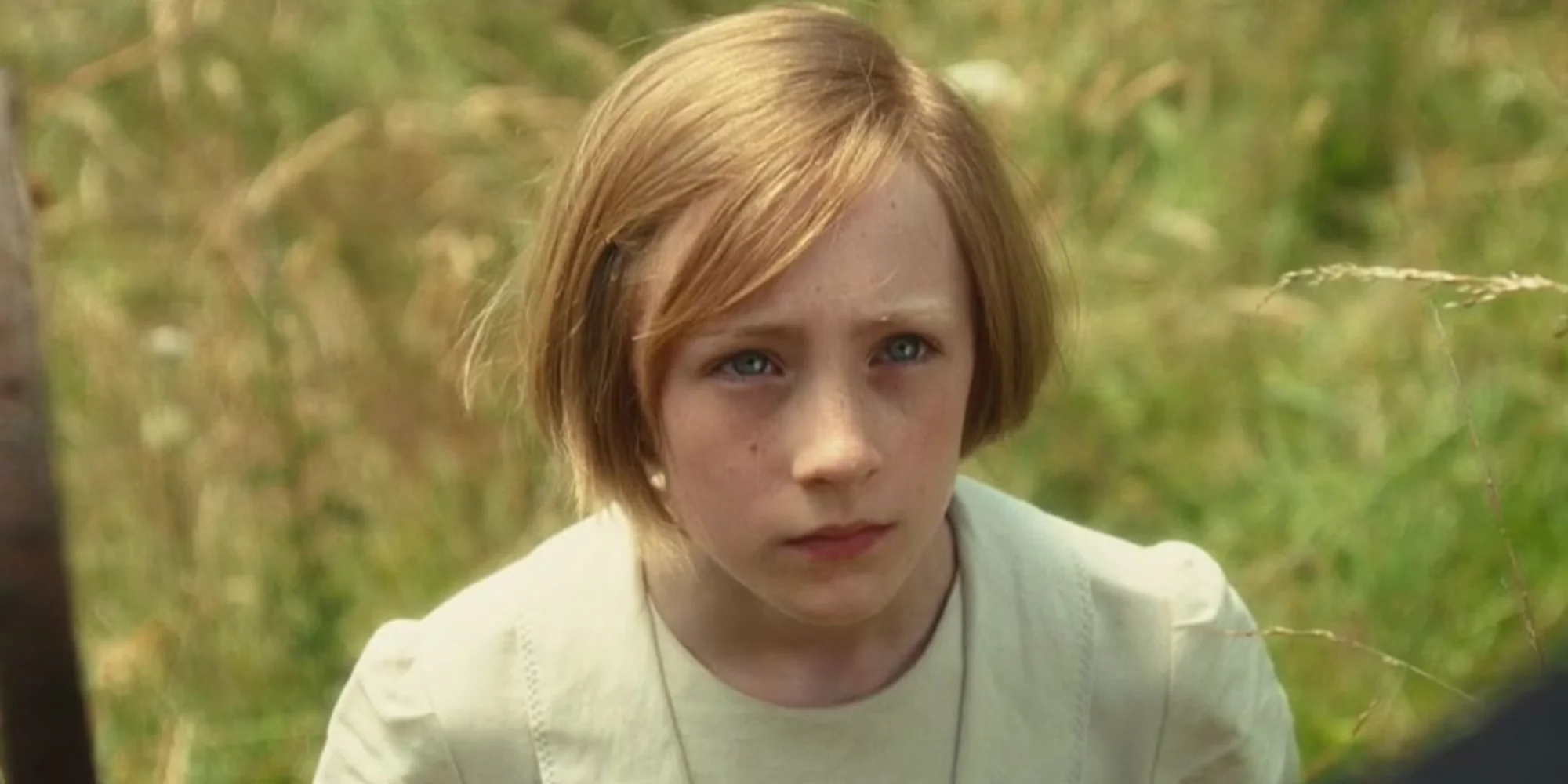 A close-up, medium shot captures a young blonde girl, portrayed by actress Saoirse Ronan as the character Briony Tallis from the 2007 film Atonement. She has short, chin-length strawberry-blonde hair with a side part and wispy bangs swept across her forehead. Her fair skin is lightly freckled, and she looks slightly off-camera to the right with wide, striking blue eyes and a solemn, unblinking expression. Her lips are softly parted in a serious, contemplative manner. She wears a simple, cream-colored short-sleeved blouse or dress with subtle stitching detail along the crew neckline and slightly puffed shoulders. The background consists of a softly blurred, sunlit green and golden grassy field, creating a shallow depth of field that keeps the focus entirely on her face.