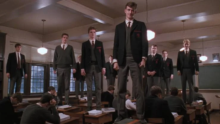 An indoor shot captures a classroom scene from the movie Dead Poets Society, where several students in school uniforms stand atop their wooden desks, while others remain seated. The room is dimly lit by hanging globe pendant lights, casting a warm glow across the wooden beams on the ceiling and the rows of desks. In the foreground, two students sit at their desks, hunched over books with their backs to the camera. Dominating the center, a student stands confidently on his desk, wearing a dark blazer over a V-neck sweater, a tie, and gray trousers, looking directly ahead with a subtle smile. To his left and right, other students stand on their desks in similar attire, looking toward the front of the room. In the background, windows with horizontal blinds reveal a dark exterior, and a large chalkboard hangs on the wall to the right. The overall atmosphere is solemn yet defiant, filled with a shared sense of quiet rebellion.