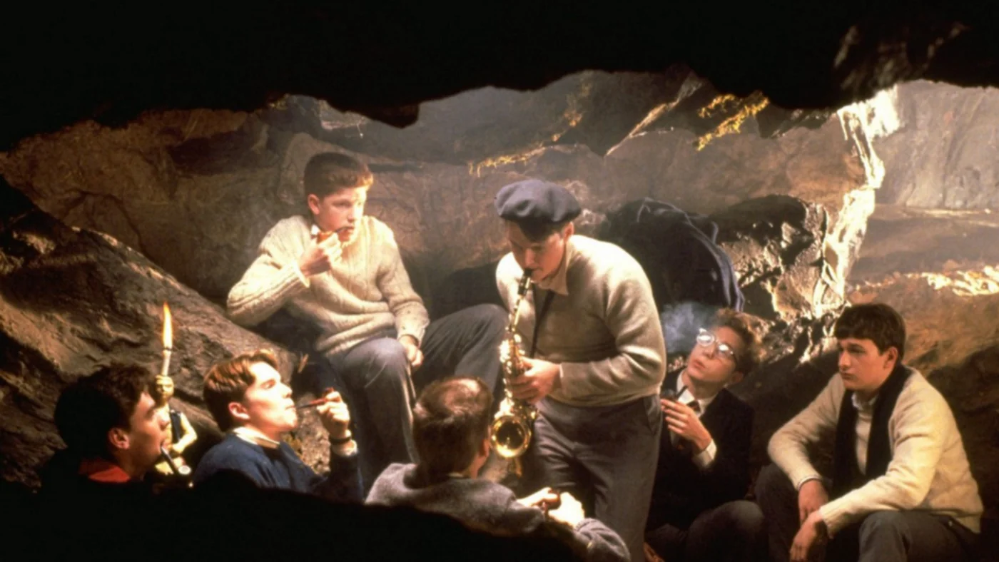 A horizontal film still captures a group of teenage boys gathered inside a dimly lit, rocky cave, lit by a soft, warm glow. The boys are dressed in 1950s-style prep school attire, including sweaters and collared shirts. In the center, a boy wearing a dark flat cap stands while playing a saxophone. Surrounding him, five other boys sit or recline against the rough stone walls of the cave; one boy on the left holds a small pipe to his mouth, while another on the right wears glasses and smokes a pipe. A lit candle rests on a small rock ledge in the lower left corner, casting light on a boy looking upward in profile. The cave opening frames the entire scene from the top and bottom with a dark silhouette.
