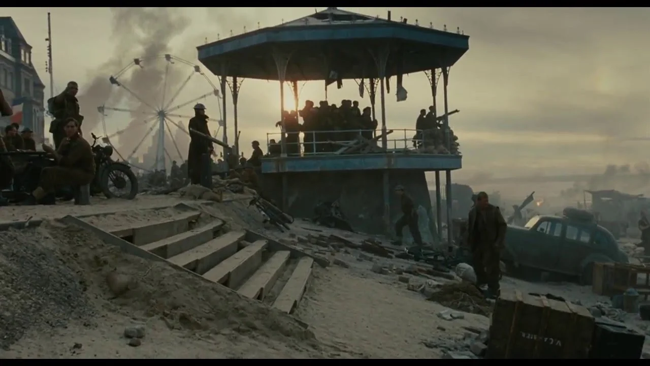 A wide shot from the 2017 war film Dunkirk shows a war-torn beachhead littered with debris, military equipment, and a vintage car, dominated by an elevated, blue-painted bandstand filled with soldiers. On the left, several soldiers are stationed near a motorcycle, while a large Ferris wheel rises into the smoke-filled sky in the background, contrasting a seaside resort with the devastation of the evacuation.