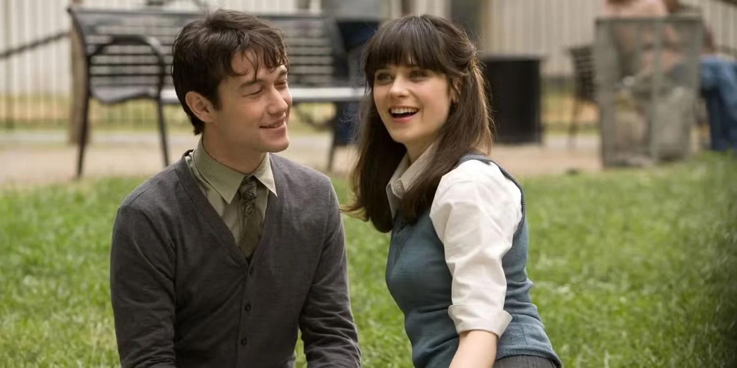 A medium, wide shot captures a young man and a young woman sitting side-by-side outdoors on a lush green lawn, looking toward the right. On the left, the man is shown in profile, facing right with a warm smile. He has short, messy dark hair and is wearing a dark grey, buttoned-up cardigan over a light olive-green collared shirt and a patterned dark tie. On the right, the woman sits with her body turned toward the right, looking forward with a cheerful, wide-open smile. She has long, dark brown hair with straight bangs, pulled back at the sides, and wears a blue sleeveless sweater vest over a white collared blouse with short sleeves. The background features a softly blurred park setting with empty wooden benches, a black trash bin, and a wire mesh fence lining a distant walkway under natural daylight.