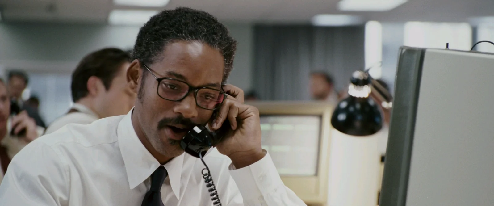 A medium close-up shot captures Will Smith portraying Chris Gardner in a brightly lit, sterile office setting. He is positioned in the center-left foreground, slightly angled toward the right. He wears rectangular, black-rimmed glasses, a white dress shirt, and a dark tie. Holding a black corded telephone receiver to his left ear, his mouth is slightly open as if speaking or reacting with intensity.The background is softly blurred, showing other office workers in white shirts, bulky vintage computer monitors, desk lamps, and beige privacy dividers typical of a late-1980s brokerage firm. A large, grey computer monitor occupies the immediate right foreground, partially framing the scene.