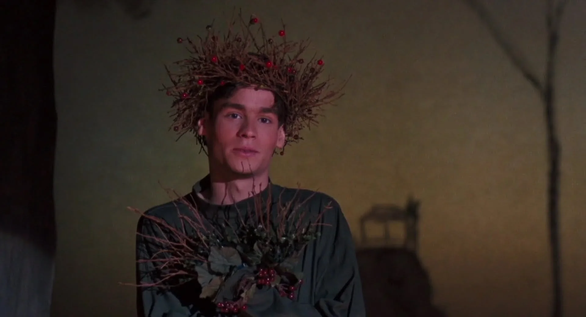 A cinematic medium close-up shot captures Neil Perry, portrayed by Robert Sean Leonard, performing on a stage under dramatic theatrical lighting. He wears a crown made of thin, tangled brown branches studded with small red berries, along with a dark green long-sleeved shirt featuring a matching bundle of twigs, leaves, and berries pinned to his chest. He gazes forward with a subtle, melancholic expression, his fair skin highlighted against a warm, softly lit backdrop painted with a hazy yellow-and-brown twilight gradient, where the faint silhouette of a bare tree and a distant structure are visible.