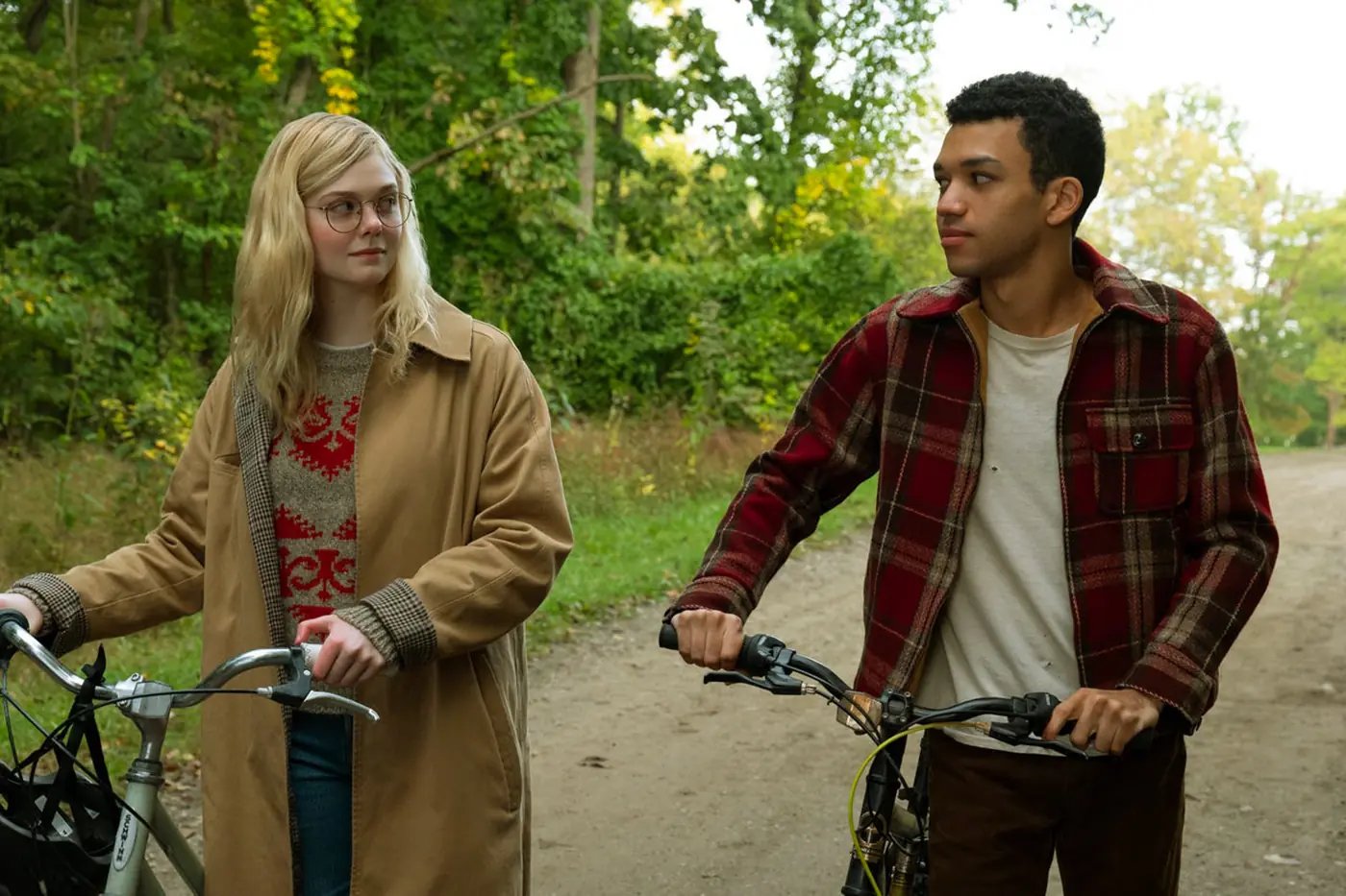 An expansive outdoor shot reveals two young adults standing with their bicycles on a dirt path running through a lush, green wooded area. On the left, a young woman with long blonde hair and round glasses looks toward the man, wearing a long beige trench coat over a textured red and white patterned sweater and blue jeans. On the right, a young man with short dark hair looks back at her, dressed in a red and brown plaid flannel jacket over a plain white t-shirt and brown pants. Both hold onto the handlebars of their classic bicycles, capturing a quiet, reflective moment surrounded by dense trees and foliage.