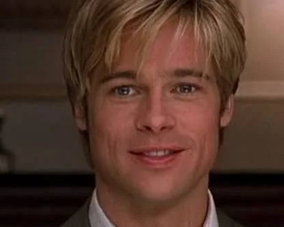 A close-up shot captures actor Brad Pitt as Joe Black, looking slightly off-camera with a gentle, contemplative expression. He has short, styled blonde hair with long bangs sweeping across his forehead, and bright blue eyes. He is dressed in a light grey or olive suit jacket over a white-and-beige striped button-up shirt, paired with a loosely knotted, muted grey patterned tie. The background is softly blurred, showing a warm, upscale indoor setting featuring a fireplace mantle, decorative items, and a glowing chandelier, creating an intimate and dramatic atmosphere.