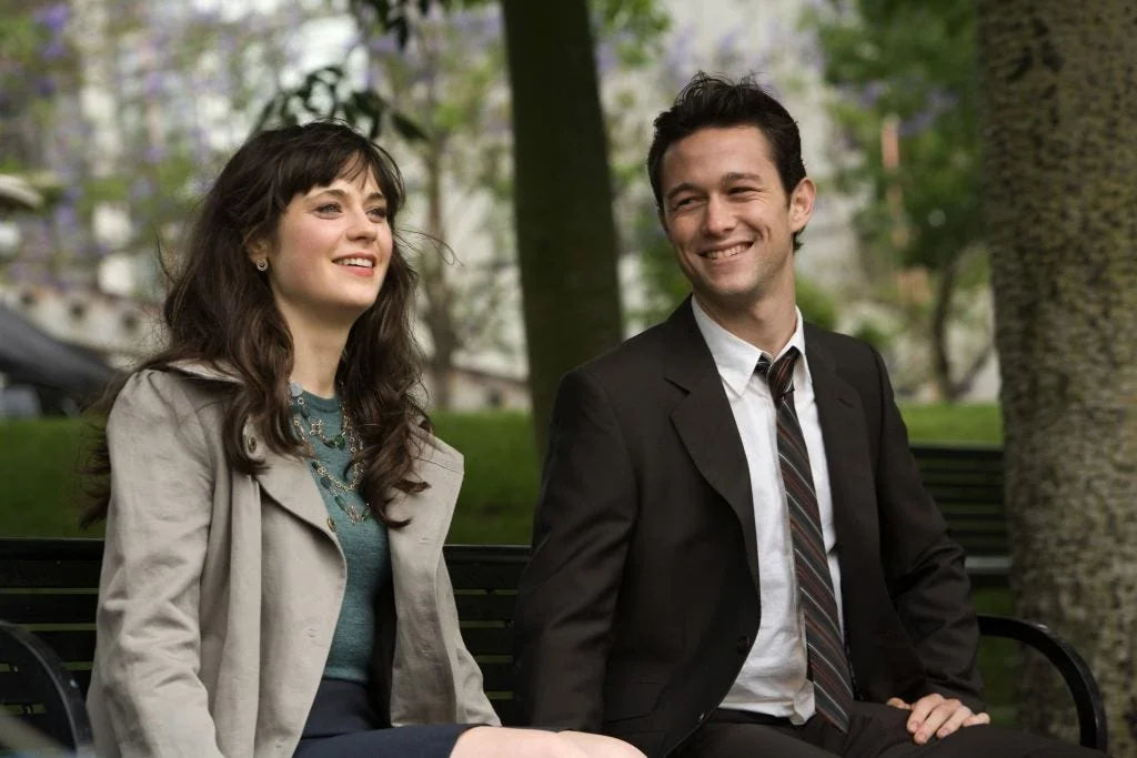 A medium shot captures a man and a woman sitting next to each other on a dark metal park bench, surrounded by a softly blurred green park landscape. The woman on the left, with long, wavy dark brown hair and full bangs, is looking slightly away to the left with a bright, genuine smile. She is wearing a light grey or beige trench-style jacket open over a teal blue top, a dark skirt, and a layered, vintage-style statement necklace with greenish beads. The man on the right sits slightly angled toward her, looking directly ahead with a relaxed, happy grin. He has short, dark, slightly spiked hair and is dressed in a dark brown suit jacket, a crisp white button-down shirt with an open collar, and a loosely tied brown, grey, and white striped tie. The background features lush green grass, trees, and hints of pale purple jacaranda blossoms out of focus, creating a bright, natural, and romantic atmosphere. A thick tree trunk frames the right edge of the shot.
