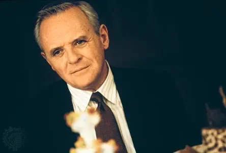 Anthony Hopkins portrays his character William Parrish in a promotional still from the 1998 romantic fantasy drama film Meet Joe Black, directed by Martin Brest. He is depicted from the chest up, positioned slightly off-center against a dark, indistinct background that suggests an elegant indoor setting. Turning his head slightly to his right, he looks directly forward with a soft, contemplative expression, his lips closed in a faint, subtle smile. He has short, neatly combed grey and white hair, clear blue eyes, and visible wrinkles around his forehead and eyes that reflect his mature age. He is dressed formally in a dark black suit jacket over a light-colored, vertically striped dress shirt, paired with a dark brown textured necktie. In the lower foreground, blurry, out-of-focus elements indicate objects on a table, including what appears to be a decorative item or a cake, though they remain heavily obscured. The lighting is warm and directional, illuminating the right side of his face while casting soft shadows on the left, enhancing the dramatic and intimate mood of the scene.