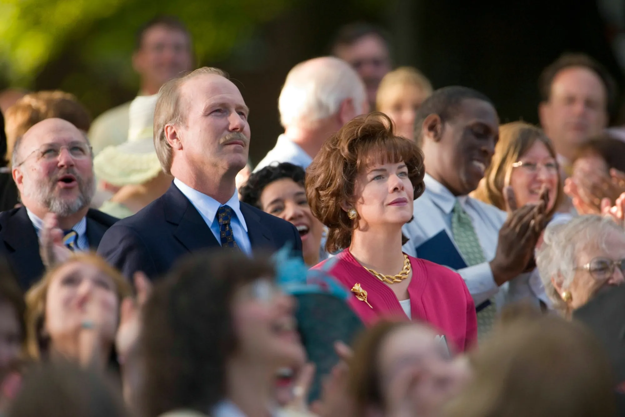 A medium shot captures a crowd of people outdoors, all looking upward and toward the right with expressions of focused attention or clapping. In the center-left stands a middle-aged Caucasian man with thinning blonde hair, a mustache, and a pensive expression, dressed in a dark blue suit jacket, white shirt, and a patterned blue tie. To his right, a Caucasian woman with voluminous, styled brown hair looks up with a serious demeanor; she wears a vibrant pink top, a thick gold chain necklace, and a gold brooch shaped like a flower. Behind them and scattered throughout the crowd are several other individuals, including a smiling Black man in a light gray suit jacket with a green tie on the right, and an older man with glasses on the far left. The background is softly blurred with hints of green foliage.