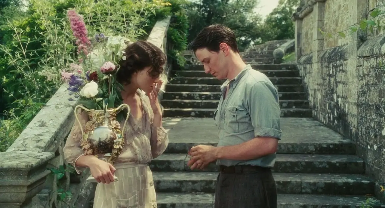 This image shows a tense interaction between two characters from the 2007 romantic war drama film Atonement. On the left, Cecilia Tallis (played by Keira Knightley) stands in a garden setting, holding a large, ornate ceramic vase filled with tall, vibrant wildflowers including purple and pink lupines. She wears a light, patterned vintage dress, her dark hair is styled in a short bob, and she holds a lit cigarette up to her mouth with her left hand while looking downward. On the right, Robbie Turner (played by James McAvoy) stands facing her on wide stone steps leading up into the background. He is dressed in a light blue, short-sleeved button-down shirt and dark trousers, looking down intently at his hands, which are held together at waist height. The backdrop features lush green foliage on the left and a weathered stone balustrade and wall on the right, capturing the lush but emotionally charged atmosphere of the English country estate scene.