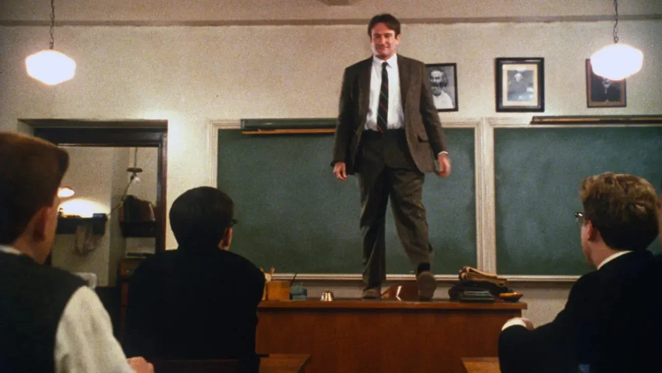 An eye-level shot captures a brightly lit classroom where a male teacher, portrayed by Robin Williams, stands confidently on top of his wooden desk. He faces a group of young male students sitting in rows before him. The teacher wears a brown suit jacket, matching trousers, a crisp white collared shirt, and a dark necktie. He smiles warmly down at his class with his hands loosely at his sides. Behind him, two large green chalkboards line the wall, topped by three small framed portraits of historical figures. Suspended from the ceiling are two glowing, white orb pendant lights that frame the scene. In the foreground, the dark silhouettes of three students face forward, their attention entirely fixed on their teacher. To the left, an open doorway reveals a glimpse into a dimly lit adjacent room.
