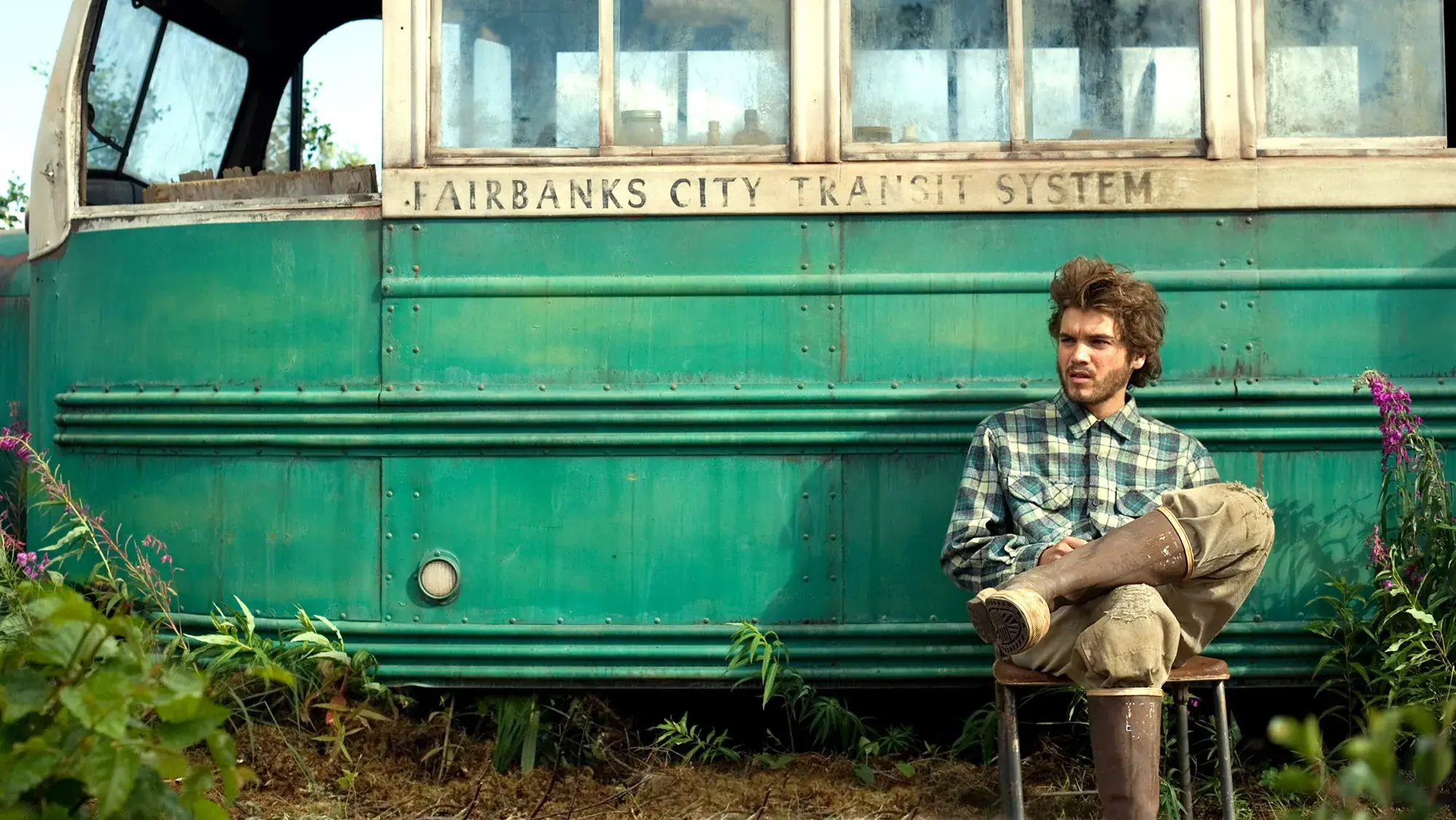A wide, eye-level shot captures a young man with messy brown hair and a beard, dressed in a green-and-white plaid button-down shirt and beige pants tucked into tall, muddy brown rain boots. He is seated on a simple wooden chair, with his legs crossed, positioned in front of a rustic, green-and-white transit bus. The side of the bus prominently features the faded stenciled text, "FAIRBANKS CITY TRANSIT SYSTEM." Lush green foliage and wild pink flowers surround the base of the vehicle, casting a serene yet isolated atmosphere over the scene.
