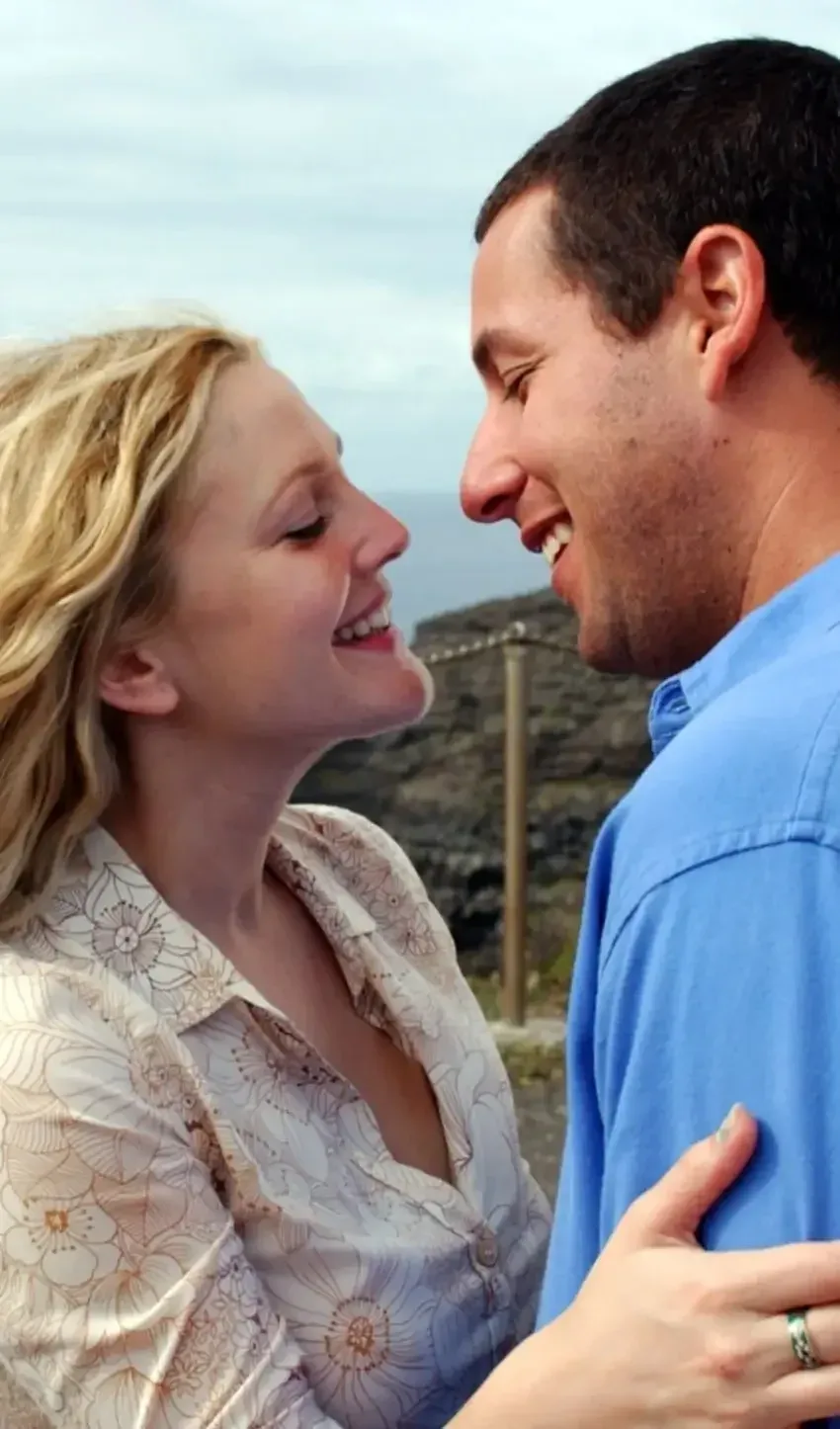 A close-up profile shot from the film 50 First Dates featuring Drew Barrymore as Lucy Whitmore and Adam Sandler as Henry Roth standing close together in an intimate embrace outdoors. Lucy, on the left, has wavy blonde hair, light skin, and a soft smile as she tilts her head upward with eyes partially closed, wearing a cream-colored collared shirt patterned with beige flower illustrations. On the right, Henry wears a blue button-down shirt and leans in with a wide, gentle grin, looking down at Lucy. A woman’s hand wearing a dark-patterned ring on her ring finger rests on Henry's lower chest/shoulder. In the background, dark coastal cliffs and a wooden railing sit against an overcast sky and soft blue ocean waters.