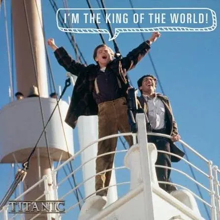 A film still from the 1997 movie Titanic captures a joyful moment at the bow of the ship against a bright, clear blue sky. Leonardo DiCaprio, portraying Jack Dawson, stands balanced at the very edge of the white prow with his arms raised wide and fists clenched in triumph. He wears a dark plaid open jacket over a purple shirt with brown suspenders and tan corduroy trousers, his mouth wide open as he shouts. A stylized white speech bubble above him reads, "I'M THE KING OF THE WORLD!". Standing just behind him to his right is Danny Nucci, playing Fabrizio De Rossi, who leans against the white metal railing. Fabrizio wears a brown coat over a white collared shirt and dark trousers, looking up with a wide, ecstatic grin. The metallic film logo "TITANIC" is positioned in the bottom left corner, while rigging lines and a large cream-colored ship mast rise into the upper left background.