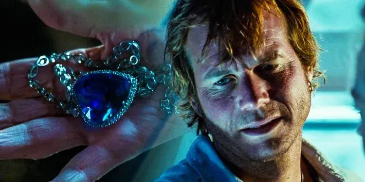 An older hand holds a large, heart-shaped blue diamond necklace—the famous Heart of the Ocean—surrounded by smaller diamonds on a silver chain, while a close-up overlay on the right shows a weathered, sun-bronzed man looking downward with a mixture of fatigue and deep contemplation. The man, a rugged treasure hunter with messy, damp brown hair, a hint of stubble, and an earring, is illuminated by an eerie, cool blue light that highlights the intense, searching look in his eyes against a blurred, clinical background.