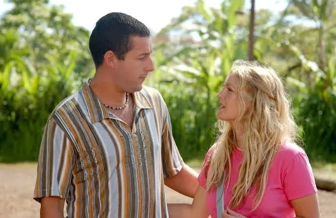 A medium close-up shot from the movie 50 First Dates captures Adam Sandler and Drew Barrymore standing outside against a blurred background of lush green tropical foliage. On the left, Adam Sandler is shown in profile, looking down toward Drew Barrymore with a serious expression. He has short dark hair and is wearing a vertically striped short-sleeved button-down shirt in shades of brown, beige, and white, paired with a beaded necklace. On the right, Drew Barrymore faces him in profile, looking up to speak. She has long, wavy blonde hair and wears a simple bright pink crewneck t-shirt with a grey bag strap across her shoulder. Her hand rests gently on his arm. The scene is brightly lit by natural daylight, casting a soft glow over both actors.