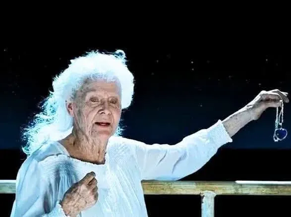 A medium shot captures elderly Rose Dawson Calvert, portrayed by actress Gloria Stuart, standing on the deck of a research vessel at night in a scene from James Cameron's 1997 film Titanic. She has long, voluminous white hair styled loosely back and wears a white, textured nightgown with smocking across the chest. She extends her left arm to the side, holding the "Heart of the Ocean" necklace—a large, heart-shaped blue diamond pendant on a silver chain—dangling delicately from her fingers over the dark ocean water. Her expression is solemn and reflective, with her lips slightly parted as she prepares to drop the precious heirloom into the sea, while her right hand is held close to her chest.