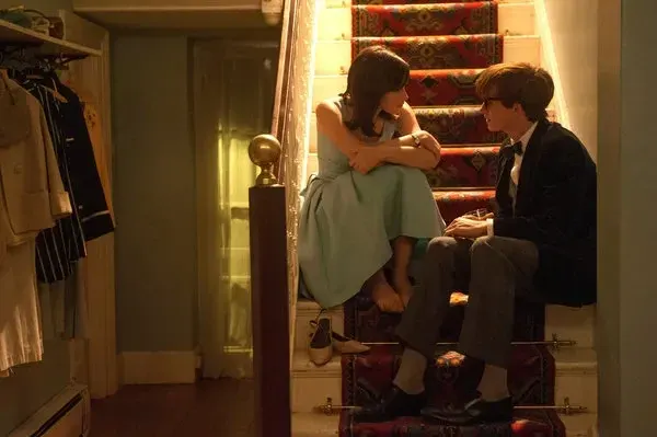 A film still captures a young woman and man sitting close together on an indoor staircase covered by a patterned red and gold runner carpet. The woman sits higher up on the left side of the stairs, wearing a sleeveless, light blue knee-length dress with full pleats. She leans forward with her arms crossed over her bare legs, looking intently at the man. Her shoes, a pair of light-colored flats, are discarded on the step below her. The man sits lower down on the right side of the staircase, wearing a dark suit jacket, gray trousers, a bow tie, and eyeglasses. He sits with his knees bent, holding a drink, and gazes back at the woman. The scene is illuminated by warm, soft lighting, with a clothing rack filled with hanging garments visible on the left side of the frame in the background.