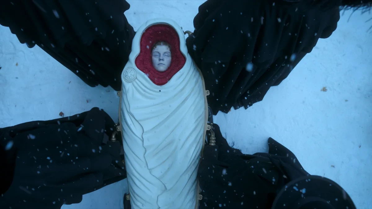 An overhead, bird's-eye view shot captures a somber funeral procession in a snowy winter setting. At the center lies a specialized, form-fitting white metallic burial capsule inspired by the historic 19th-century Fisk design. The face of a deceased woman, Claire Frankenstein (played by Mia Goth), is visible through a small glass viewing pane surrounded by a textured, deep crimson velvet lining. Flanking the white coffin, pallbearers clad in heavy black mourning cloaks and wide-brimmed hats carry the body through a light snowfall, creating a high-contrast cross formation against the snow-covered ground.