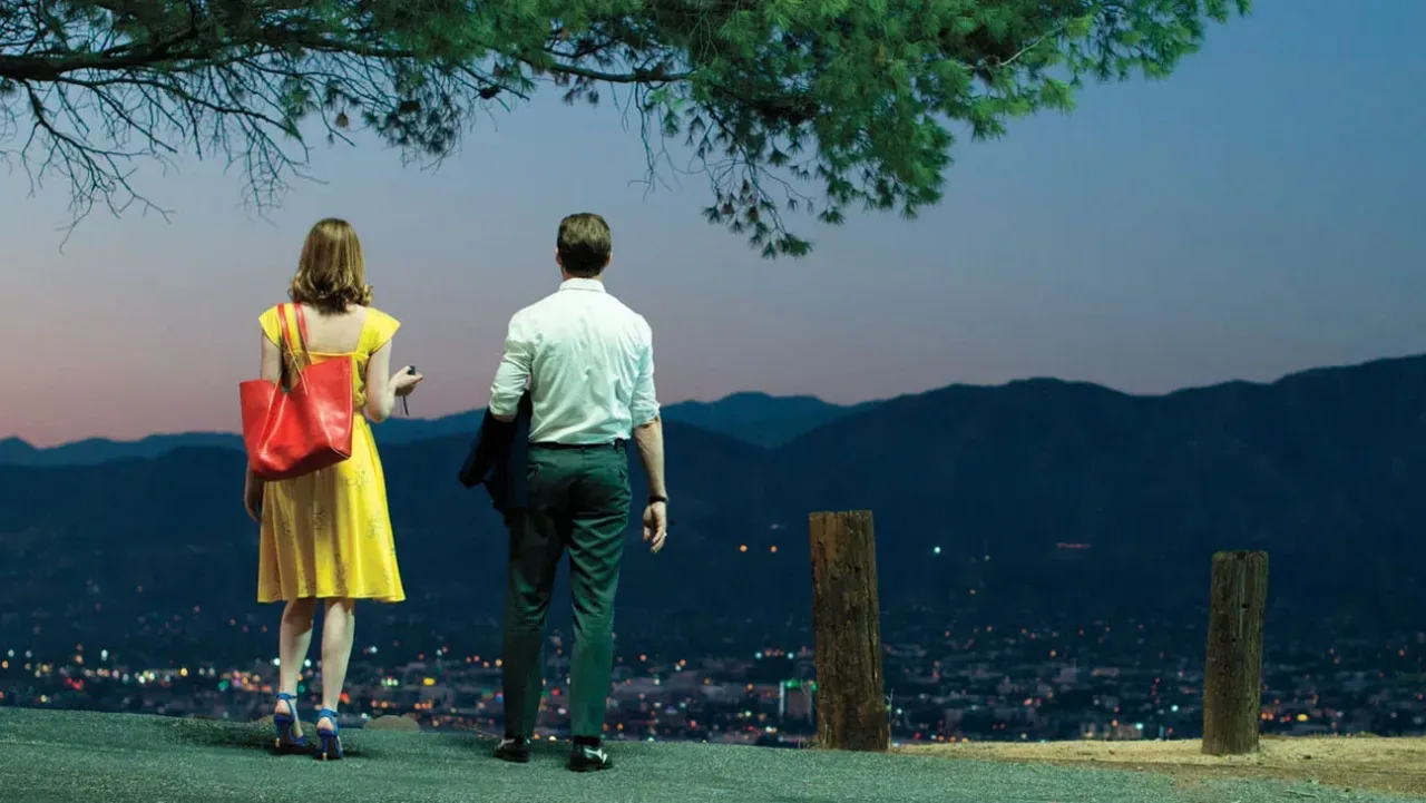 A wide shot captures a woman and a man standing side-by-side from behind, overlooking a sprawling city valley at twilight from a high vantage point. The woman on the left wears a vibrant yellow A-line dress, bright blue heeled sandals, and carries a large red leather tote bag over her right shoulder. The man on the right wears a crisp white button-down shirt, dark green tailored trousers, and dark dress shoes, holding a dark jacket over his right arm. They stand on a paved overlook lined by short wooden posts on the right. In the background, rolling blue mountain ridges silhouette against a soft dusk sky fading from orange near the horizon to dark blue above. Below the mountains, the city lights begin to twinkle across the valley floor. A leafy tree branch frames the upper edge of the scene from the top center.