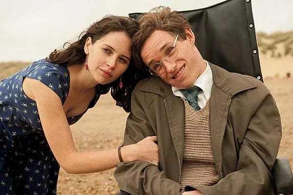 In this promotional film still, actors Eddie Redmayne and Felicity Jones lean close together against an outdoor, sandy backdrop. On the right, Redmayne portrays Stephen Hawking, sitting in a black-backed wheelchair with his head tilted sharply to the left. He wears thin, wire-rimmed glasses, a slightly crooked grin, a white collared shirt with a green patterned tie, a tan knit sweater vest, and an oversized brown trench coat. Leaning heavily against his right shoulder from the left side of the frame is Jones, portraying Jane Wilde Hawking. She rests her head against his, gazing forward with a gentle, soft expression. Her long brown hair falls loosely around her face, and she wears dangling red earrings paired with a dark blue short-sleeved dress covered in a small yellow floral pattern.