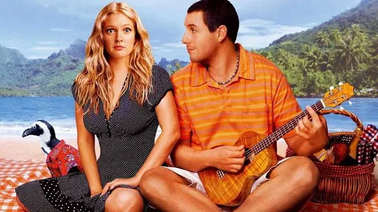 A wide promotional studio photograph for the romantic comedy film 50 First Dates features actors Drew Barrymore and Adam Sandler sitting cross-legged on a red-and-white checkered picnic blanket on a sandy beach. On the left, Drew Barrymore sits upright, facing forward with a wide-eyed, surprised, and slightly confused facial expression. She has long, wavy blonde hair parted down the middle and wears a short-sleeved, black V-neck dress with white polka dots. On the right, Adam Sandler is turned in profile toward Barrymore, looking at her with a gentle, smiling expression. He has short dark hair and wears an orange and yellow striped polo shirt, white shorts, and a dark beaded necklace while holding and strumming a small acoustic ukulele. Behind Barrymore's right hip, a small penguin wearing a red Hawaiian shirt stands on the blanket, looking towards the left. To the right of Sandler sits a woven wicker picnic basket filled with items, including a bottle. The background depicts a tropical coastline with calm blue ocean water, a distant shoreline, and lush, green, tree-covered mountains rising into a light blue, hazy sky.