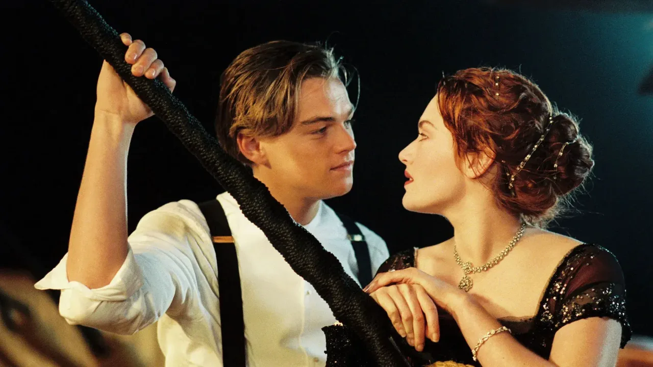 A medium shot captures Jack Dawson, played by Leonardo DiCaprio, and Rose DeWitt Bukater, played by Kate Winslet, looking intensely into each other's eyes against a dark night background on the deck of the ship. On the left, Jack is depicted from the waist up with light brown, side-parted hair with a few strands falling over his forehead, wearing a white button-down shirt with rolled-up sleeves and black suspenders. His right arm is raised, gripping a thick, dark, textured rope or post that slices diagonally through the frame. On the right, Rose is seen in profile from the chest up, her red hair styled in an elegant updo secured with a silver jeweled hairpiece. She wears an exquisite black evening gown with sheer cap sleeves detailed with glittering beadwork, a delicate silver rhinestone collar necklace, and a matching bracelet on her left wrist, which rests gently near Jack. The lighting highlights their facial features and creates a romantic, intimate atmosphere.