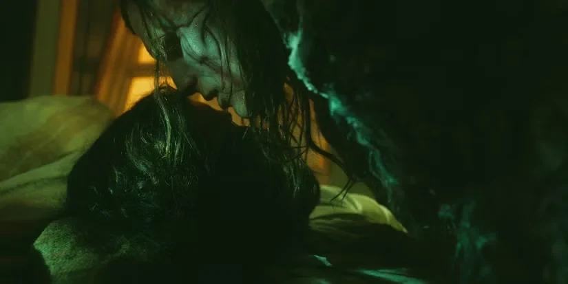A dimly lit, close-up shot bathed in an eerie green glow captures an intimate and tragic moment between two figures. From the top of the frame, a gaunt, heavily textured creature with long, damp strands of hair hanging over its pale face leans down toward a man lying flat on his back. The man beneath, featuring dark, tousled hair and a beard, looks upward as the creature moves close enough for their foreheads and noses to gently touch in a tender gesture. A heavy, dark, textured fabric shadows the right side of the composition, while a faint, warm yellow light cuts through the background on the left, casting a stark contrast against the dominant emerald shadows.