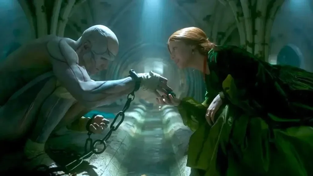 A cinematic medium shot depicts a tense, intimate interaction between a heavily mutated, muscular, and hairless creature crouching on the left and a woman in an elegant period dress kneeling on the right. The scene takes place within a dark, gothic, stone-arched subterranean dungeon or drainage corridor with pools of water reflecting light along the center floor. The pale, alabaster-skinned creature is muscular yet gaunt, with geometric, phrenology-like lines across its bald head. Heavy black chains encircle its wrists, trailing down to the stone floor. Reaching forward, the creature gently touches hands with the woman, who leans in with a look of intense, quiet curiosity. She has long red hair tied back and wears an elaborate, voluminous emerald-green gown. Overhead, intense, dramatic teal and white spotlights pierce down through the gloom, casting stark shadows and illuminating the heavy, humid atmosphere between them.