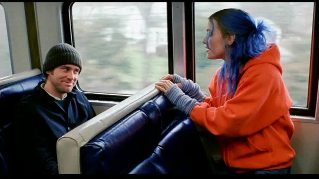 A film still captures a side-profile view of a young woman with vibrant, messy blue hair tied back, leaning over the back of a blue vinyl train seat to talk to a man. She is positioned on the right side of the frame, wearing a bright orange zippered hoodie with grey-and-white striped knit arm warmers pulled over her hands. Her expression is animated as she speaks. On the left, a man sits facing her, leaning against the window side of the blue bench seat. He wears a dark knit beanie pulled low over his forehead, a white undershirt, and a dark jacket. He looks up at her with a gentle, slight smile. Behind them, large rectangular train windows reveal a blurred, moving green and white landscape outside, indicating the train is in motion.