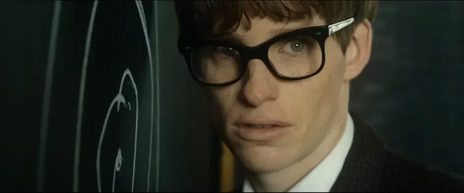 An actor portrays a young Stephen Hawking, wearing thick, black-rimmed glasses and a tweed jacket, staring intently with an expression of sudden realization or deep concentration next to a chalkboard covered in white chalk diagrams. Close-up of Eddie Redmayne in The Theory of Everything.