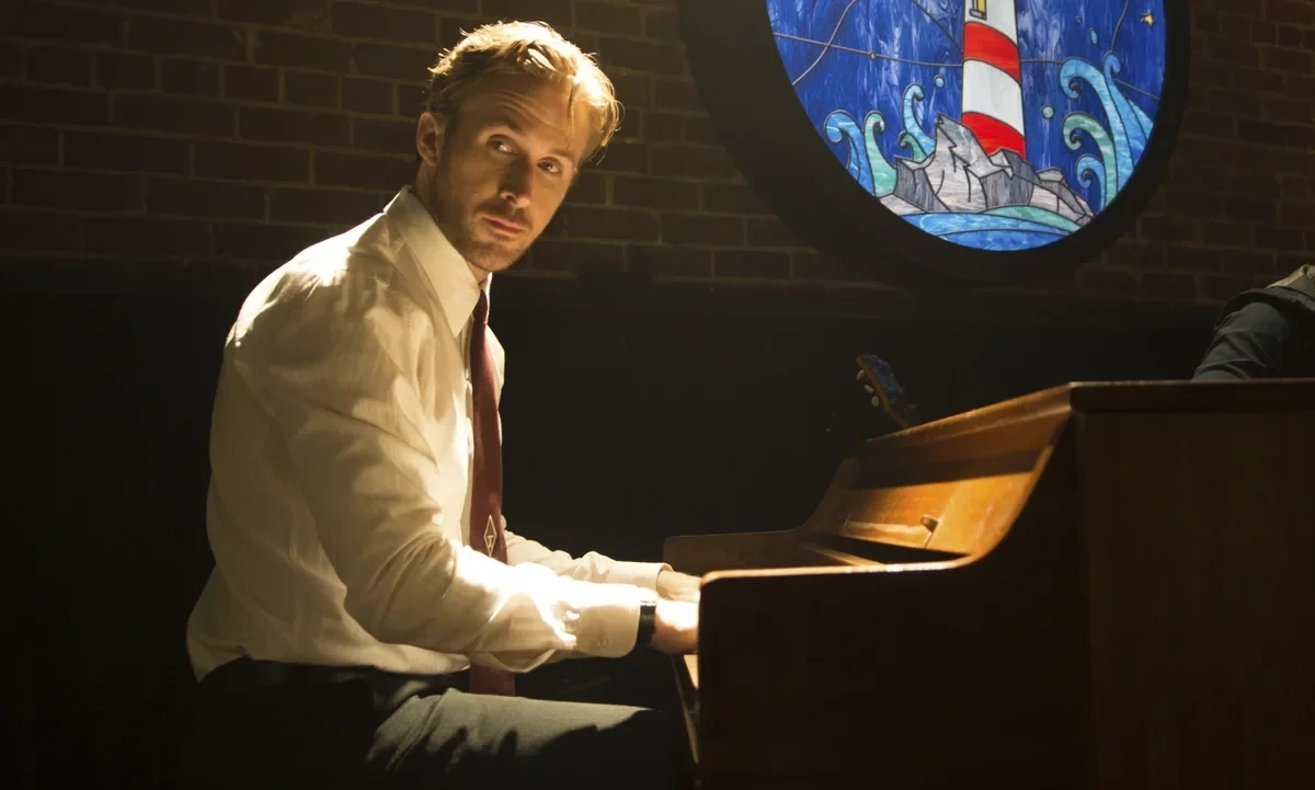 An indoor eye-level long-shot captures Sebastian (Ryan Gosling) in a white dress shirt and dark pants, seated at a brown upright piano in a restaurant with brick walls, playing the piano as he looks over his right shoulder in profile. Sebastian sits with his back straight and hands on the piano, looking back over his right shoulder with an intense gaze. His blonde-brown hair is styled neatly, and he wears a white collared dress shirt with the sleeves buttoned at the wrists, dark pants, and a thin brown tie. The upright piano is brown and has a matte finish. The brick wall behind Sebastian is a warm brown color, and a round stained-glass window featuring a lighthouse on rocks amidst blue waves is positioned above the piano. The lighting is warm and directional, casting a soft glow on Sebastian and highlighting the textures of the brick and piano. The atmosphere appears to be a dimly lit jazz club or restaurant, creating a moody and intimate setting.