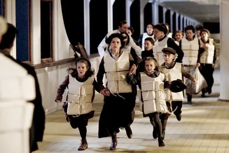 A group of actors, including a mother and three children wearing early 20th-century clothing and bulky white canvas life jackets, run in a panic down the illuminated outdoor wooden deck of a large ship at night.