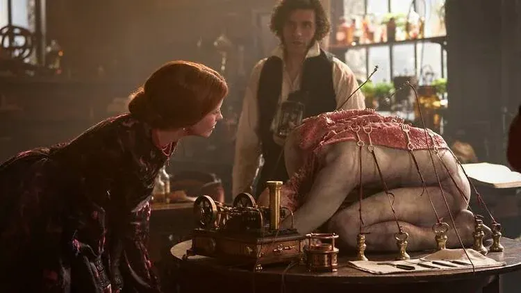 A cinematic medium shot from Guillermo del Toro's gothic film adaptation captures a dimly lit, amber-toned laboratory where three figures gather around a dark wooden table. In the center, a pale human-like creature with visible muscle definitions is hunched over on its hands and knees, facing away from the camera. Its back is covered with a textured, raw pink tissue or meat-like layer, integrated with fine wires and small brass or gold metallic clamps that run down to a series of tiny weighted stands on the table surface. On the left side of the frame, a woman with auburn hair pinned up in a classical style, wearing a dark Victorian gown with intricate patterns, leans in with intense curiosity to closely inspect the creature. In the background, positioned between the two figures, stands a man with dark curly hair and a mustache, dressed in a white linen shirt, a dark vest, and red-trimmed gloves. He holds a small scientific object in his hand, observing the scene with a focused expression. The room is hazy with soft, diffused sunlight filtering through a grid window pane in the upper left, casting a warm glow across intricate machinery, small glass vials, and open books cluttering the workspace.