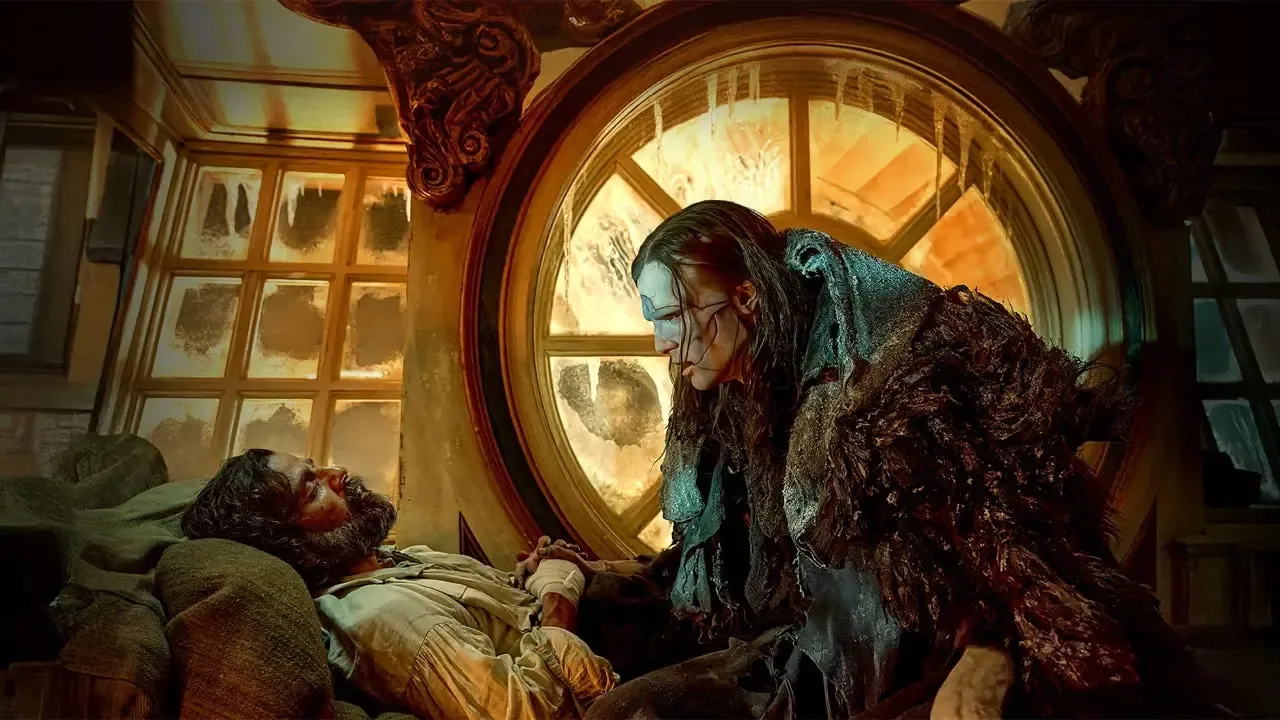 A dramatic cinematic still depicts two central characters inside a dimly lit wooden ship cabin under an ominous atmosphere. On the left, a pale, heavily bearded man, Oscar Isaac as Dr. Victor Frankenstein, lies sickly on his back upon a pile of canvas pillows with his hand wrapped in bandages. Standing over him on the right is Jacob Elordi as the towering, gaunt Creature, featuring visible surgical stitching across his pale face and dark stringy hair, draped in a massive, textured dark fur and seaweed cloak. The Creature leans slightly forward, holding Victor's bandaged hand in a tense, intimate moment of final reconciliation. They are framed by a large circular multi-paned porthole window in the center background that glows with a cold, warm amber light and is frosted with long icicles hanging down from its top rim.