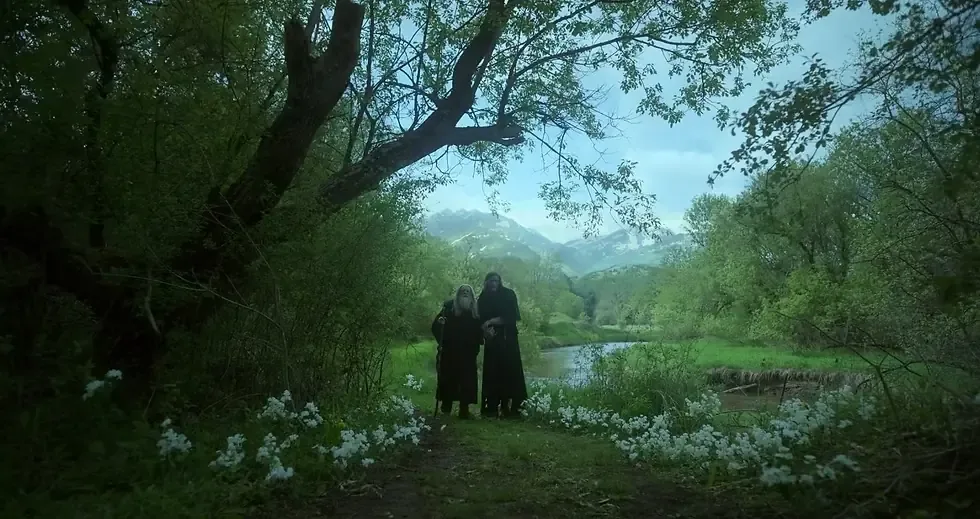 A wide, cinematic shot of a vibrant green landscape under a pale, overcast sky. In the foreground, a grassy dirt path is lined with clusters of delicate white wildflowers. Two figures wearing long, dark hooded cloaks walk side-by-side along the path toward the center of the frame. One figure appears older with long gray hair and a beard, holding a walking stick. Dense, leafy green trees and overhanging branches frame the upper and outer edges of the scene. In the background, a narrow river winds through a grassy meadow, leading toward hazy, distant mountains. The overall atmosphere is serene and moody, with a cool blue and green color palette.
