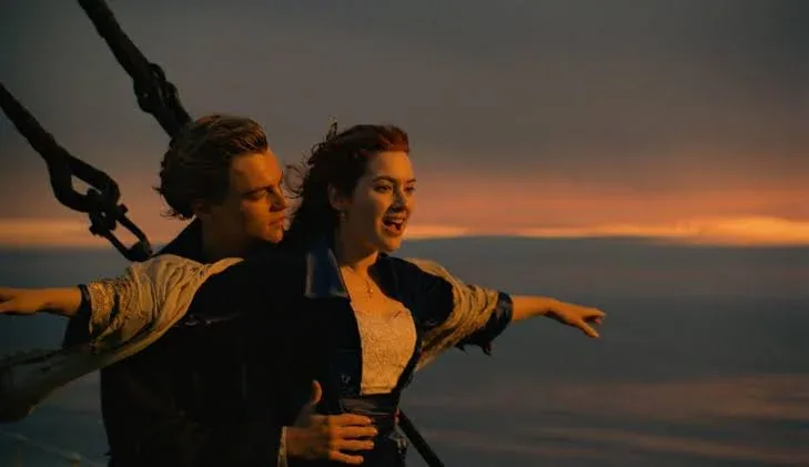 A cinematic medium shot from the 1997 film Titanic featuring Jack Dawson (played by Leonardo DiCaprio) and Rose DeWitt Bukater (played by Kate Winslet) at the bow of the ship during a vibrant orange and golden sunset over the ocean. Rose stands at the very edge with her arms extended wide, smiling with open-mouthed awe and exhilaration. Her red hair blows back in the wind, and she wears a dark blue velvet coat over a white lace dress. Jack stands closely behind her, holding her securely by her waist with one hand while stretching his other arm out parallel to hers. He looks at her affectionately with a soft smile. Large, dark metal rigging ropes and cables anchor into the frame on the left, cutting diagonally against the glowing horizon line where the orange sunset meets the dark, calm sea below.