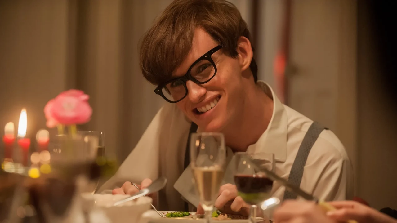 A medium shot captures actor Eddie Redmayne portraying Stephen Hawking in the film The Theory of Everything, seated at a warmly lit dining table. He is leaning slightly forward and tilting his head with a wide, bright smile while wearing thick, black-rimmed glasses and a white collared shirt with grey suspenders. In the softly blurred foreground, lit candles, a pink flower, and glasses filled with white and red wine are scattered across the table, adding an intimate, cozy atmosphere to the scene.