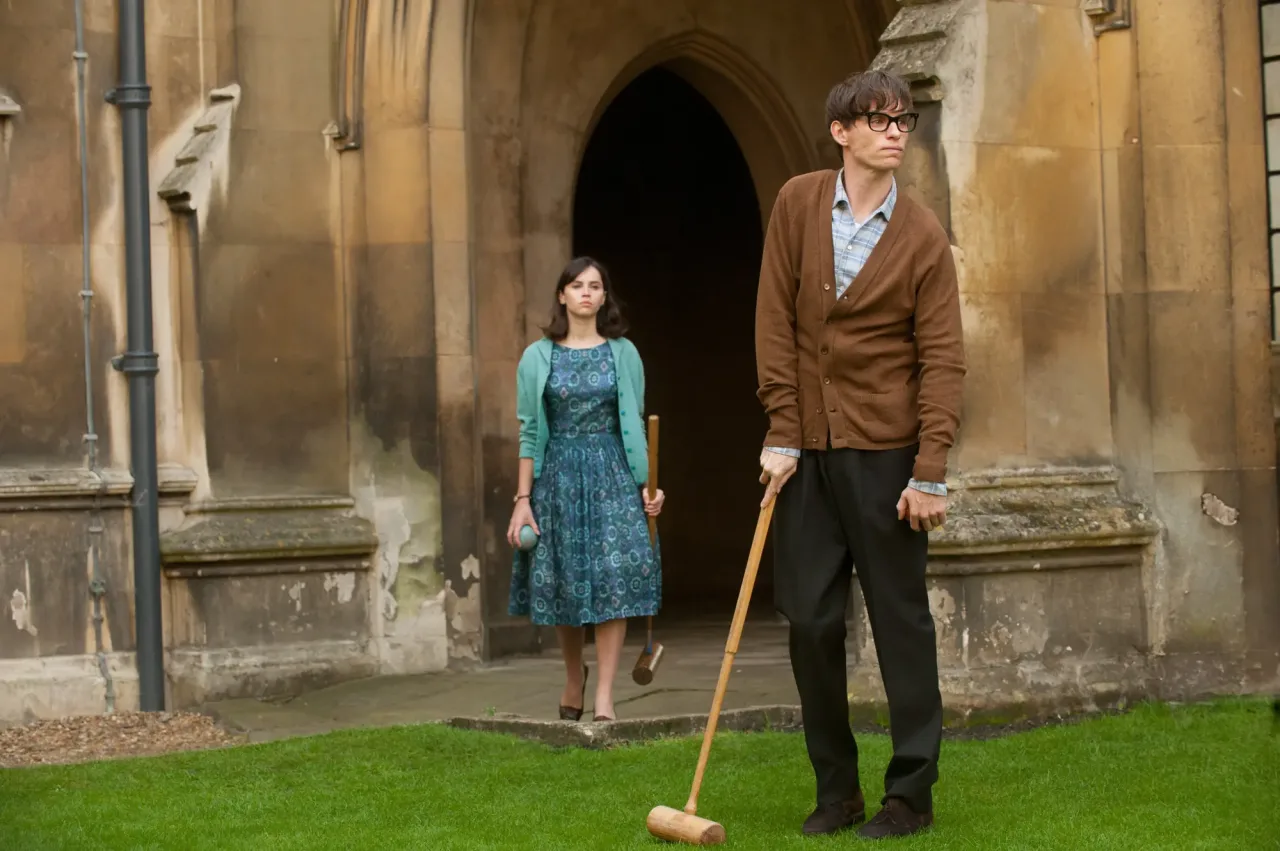 A wide, eye-level shot captures a man and a woman in mid-20th-century attire standing on a manicured green lawn in front of a weathered stone building with a Gothic-style arched doorway. The man, with messy dark hair and black-framed glasses, stands on the right, leaning slightly forward as he holds a wooden croquet mallet resting on the grass. He wears a brown knit cardigan over a light-colored plaid button-down shirt and loose-fitting dark trousers. His expression is contemplative, looking slightly off-camera to the right. A few paces behind him and to the left, a woman stands near the arched entryway, holding a croquet mallet in one hand and a blue croquet ball in the other. She wears a patterned teal knee-length dress under a matching teal cardigan, with her dark hair styled in a shoulder-length bob. The background features aged, tan stone walls of an academic or historic building, complete with a dark downpipe on the far left, creating a scholarly and vintage atmosphere.