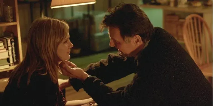 An indoor, dimly lit, close-up profile shot captures a tender moment between Mary Svevo (Kirsten Dunst) and Dr. Howard Mierzwiak (Tom Wilkinson) from the film Eternal Sunshine of the Spotless Mind. Mary sits on the left, looking up softly at Howard, who leans in closely from the right. He gently holds her chin with his left hand as they gaze into each other's eyes. The warm glow of a lamp overhead illuminates the scene, highlighting bookshelves on the left and a wooden chair in the background.