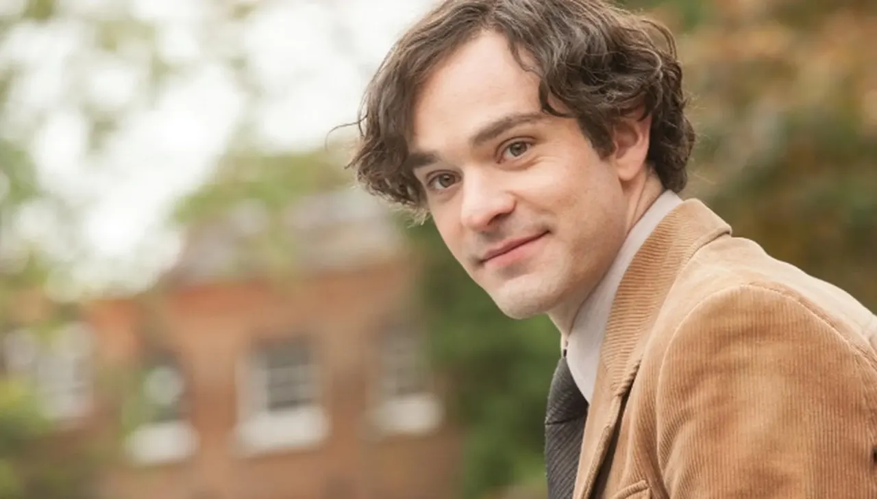 A close-up shot of English actor Charlie Cox portraying Jonathan Hellyer Jones in a scene from the 2014 biographical drama film The Theory of Everything. He has wavy brown hair, is looking directly at the camera with a gentle, subtle smile, and is dressed in a tan corduroy blazer, a white collared shirt, and a dark patterned necktie. The background shows a soft, shallow depth-of-field blur of an outdoor brick building and leafy green trees.