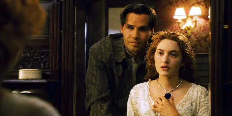 A medium cinematic shot from the 1997 film Titanic featuring two characters standing inside a wood-paneled stateroom. On the right, Rose DeWitt Bukater, played by Kate Winslet, is depicted from the waist up. She has fair skin, wavy reddish-brown hair parted down the middle, and a look of quiet apprehension. She wears a white, delicate lace-trimmed dress and a prominent white gold or platinum necklace featuring a large, heart-shaped blue diamond pendant—the Heart of the Ocean—which she holds lightly with her right hand. Standing closely behind her shoulder is Caledon "Cal" Hockley, played by Billy Zane. He is a clean-shaven man with dark, slicked-back hair, looking directly toward the left side of the frame with a subtle, confident smile. He wears a dark, patterned grey coat over a high-collared light shirt. The background consists of rich, dark wood doors and paneling, with an ornate gold sconce lamp glowing softly against a red patterned wallpaper on the right. In the blurred foreground on the far left, the back of another character's head is partially visible.
